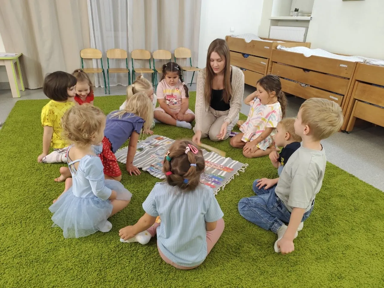 Ранкове коло в укритті садочка Unique Kids у Чайках: діти з педагогинею на зеленому килимі, позаду ліжка для денного сну