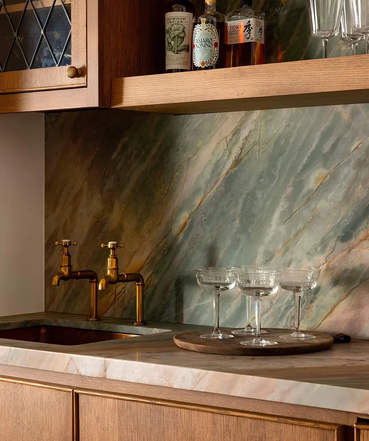 A custom wet bar with a green stone backsplash, brass faucets and wood cabinetry